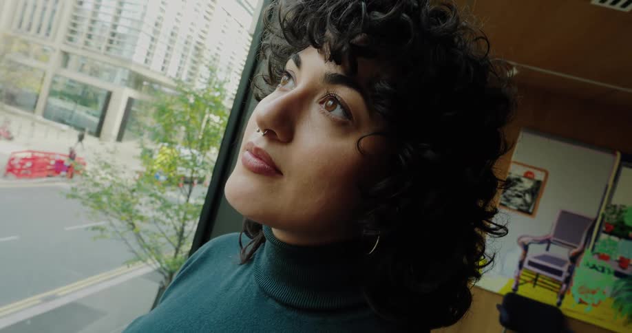 A woman with curly dark hair and a nose piercing stands by a large window in a modern office gazing out at the busy city street below she holds a small portable fan as she looks up seemingly enjoying a moment of reflection