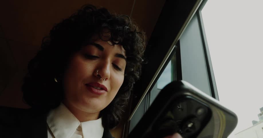 The video shows a closeup of a woman with dark curly hair and a nose ring she is looking at her phone which she is holding up near a large window the natural light from the window illuminates her face as she smiles and interacts with the content on h