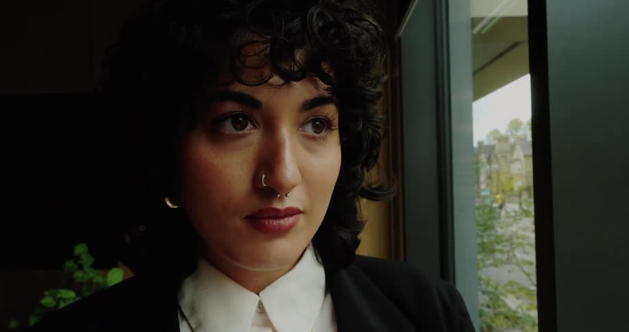 A professional woman with dark curly hair dressed in a black blazer and white shirt initially looks down at her phone by a large window then slowly raises her head puts the phone down and looks thoughtfully out the window