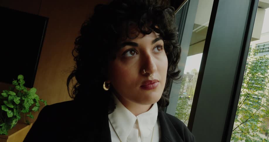 The video features a woman with dark curly hair dressed in a black blazer and white collared shirt standing near a large window she is looking out pensively at the green trees and buildings visible outside with her expression suggesting contemplation