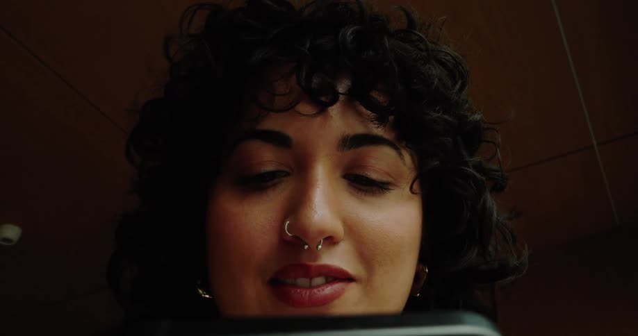 The video shows a closeup lowangle shot of a woman with dark curly hair looking down at a smartphone she is holding she has a nose ring and a septum piercingshe occasionally glances up from the phone and her mouth moves slightly as if she is quiet