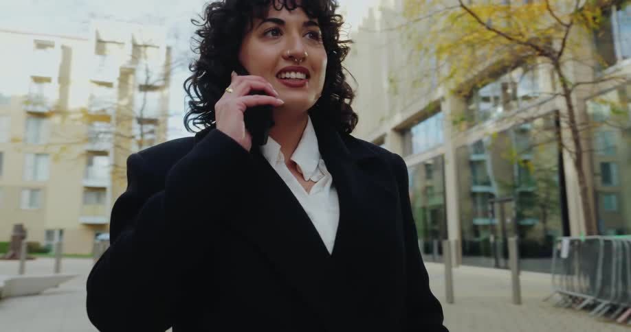 A woman with dark curly hair and wearing a black coat over a white shirt is walking outdoors in an urban area while talking on a cellphoneshe appears engaged and smiles as she speaks with modern buildings and a tree with yellow foliage visible in the