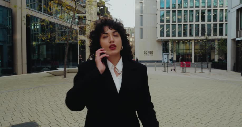 The video shows a medium shot of a woman in a black suit and white shirt walking quickly through a modern office complex plaza she is initially looking at her phone before bringing it to her ear to answer a call