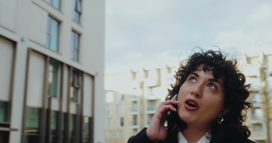 The video is a closeup shot of a woman with curly hair wearing a black blazer and white shirt speaking on a cell phone in an urban setting she looks up and around suggesting she is trying to locate something or someone while on the call