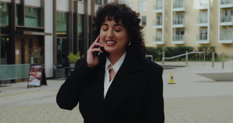 A woman with curly hair smiles while talking on her cell phone as she walks outside a modern building