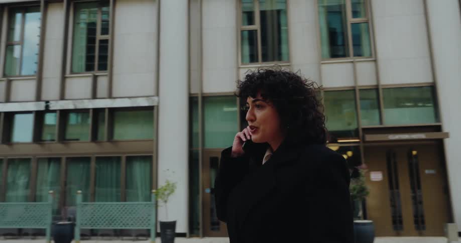 A woman with curly dark hair dressed in a black coat and white shirt is walking out of a hotel entrance clayton hotel while talking on her phone she walks past the camera and continues away from the building towards a more open area