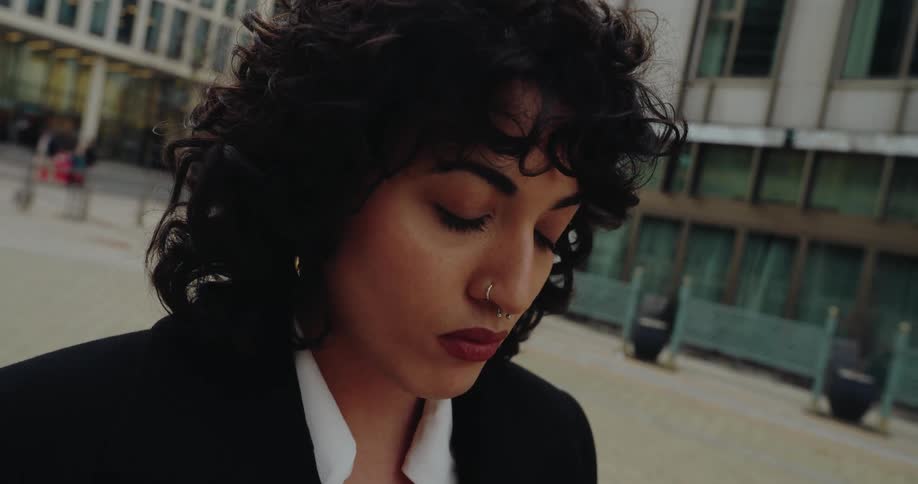 The video is a closeup tracking shot of a woman with curly hair walking outdoors in a city settingshe is looking down at her phone as she walks past modern office buildings