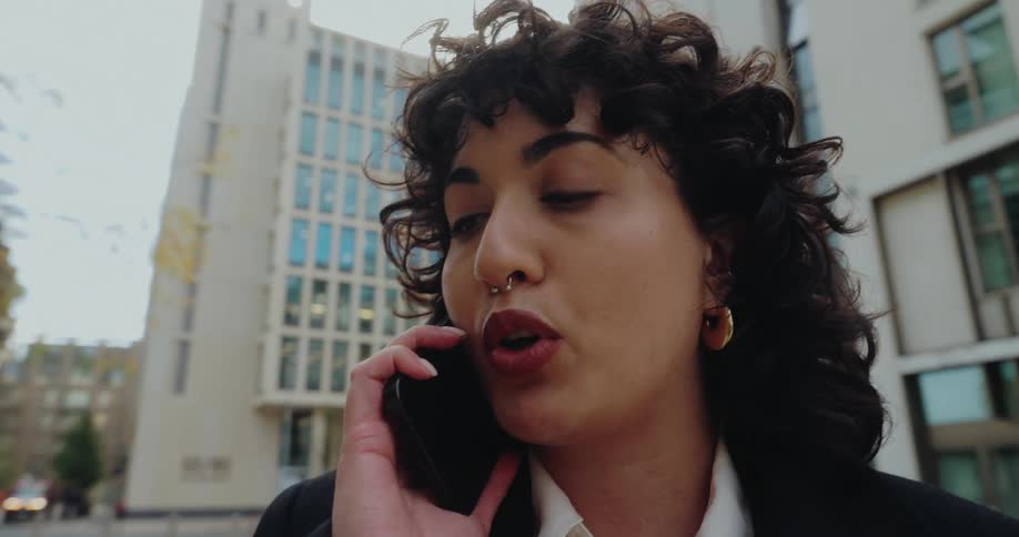 A close low angle view of a young woman with curly hair and a nose ring speaking intensely on a mobile phone outside near tall city buildings