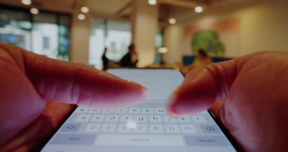 A close view of a persons hands typing a message on a smartphone keyboard in a brightly lit indoor space with blurred people in the background