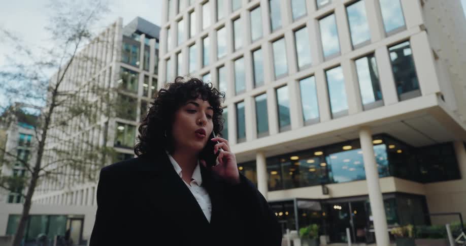 A woman with dark curly hair and wearing a black blazer walks outside talking animatedly on a cellphoneshe is framed in a lowangle shot against a modern multistory office or commercial building with large repeating windows