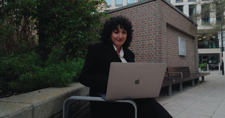 A woman is sitting outside a city building typing on her computer