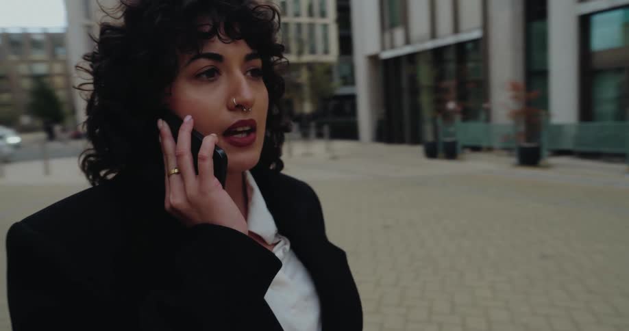 This video features a closeup of a young woman with curly hair a nose ring and a gold ring on her finger walking outside while talking on her smartphoneshe is dressed in a dark blazer over a white shirt with modern office buildings visible in the