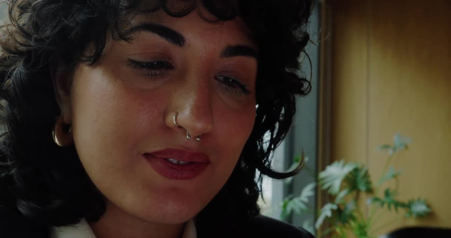 This is an extreme closeup video of a woman looking down at a smartphone she is holding she has curly hair a septum piercing and appears to be smiling slightly or reacting positively to content on the screen