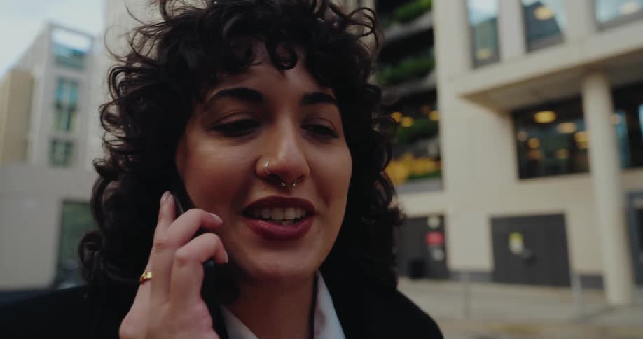 A happy woman in professional black attire is looking forward while talking on a phone outside a modern office building