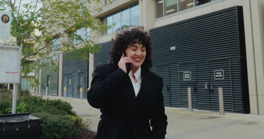 A woman with dark curly hair wearing a black coat over a white collared shirt is walking outdoors near a modern buildingshe answers a call on her phone and smiles while talking continuing to walk down the sidewalk