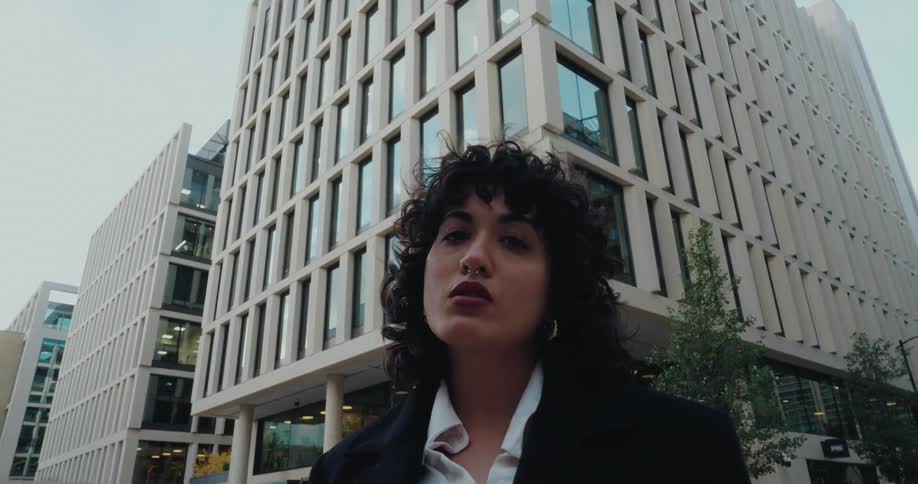 Young adult woman wearing a black blazer and a white blouse standing on the streets of a metropolitan area outdoors during day time