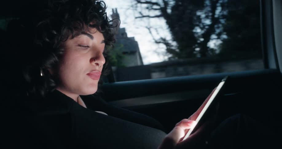 The video features a woman with curly hair sitting in the back seat of a vehicle illuminated primarily by the glow of the tablet she is holding she is looking intently at the screen occasionally licking her lips in concentration