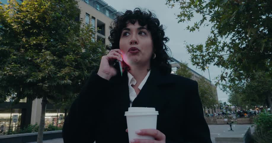 A young woman is holding a white cup and talking on a phone that is to her ear in an outdoor setting