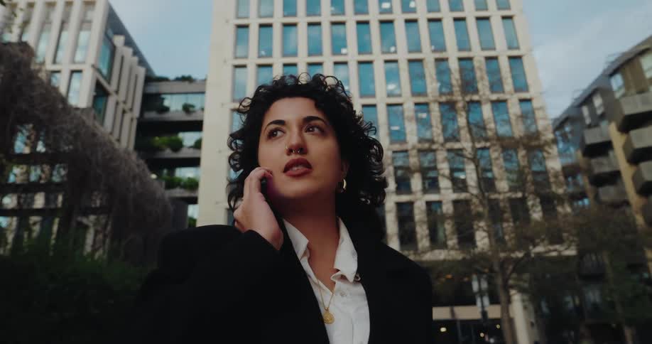 A young woman with long curly hair dressed in a suit answers a phone call in the middle of the street at midday