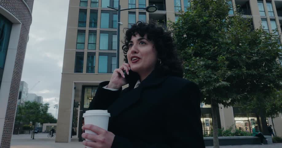 A smiling woman with curly hair walks through a city chatting on her phone and holding a coffee cup