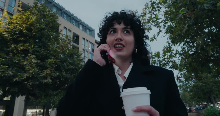 A young woman stands in the middle of the street holding a disposable coffee cup while answering a phone call