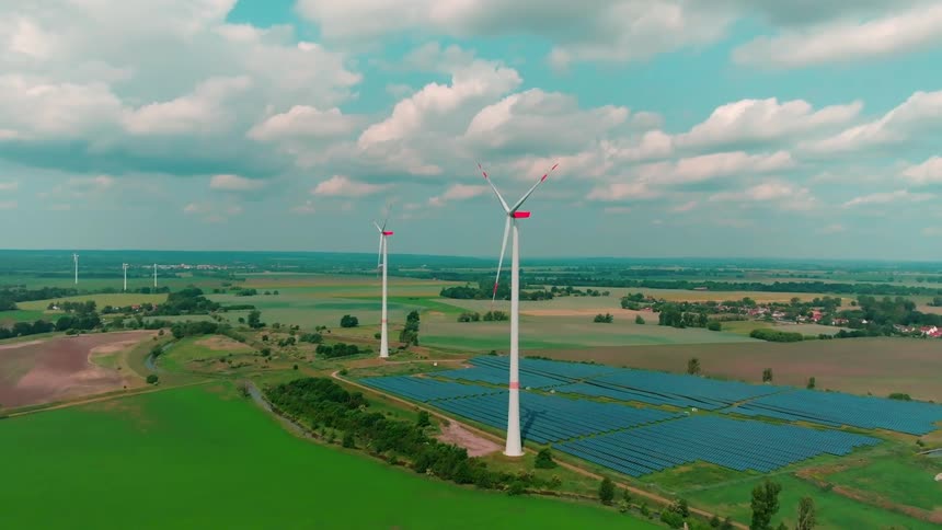 A highangle aerial view overlooks white wind turbines spinning above green fields and a solar farm under a blue sky with clouds