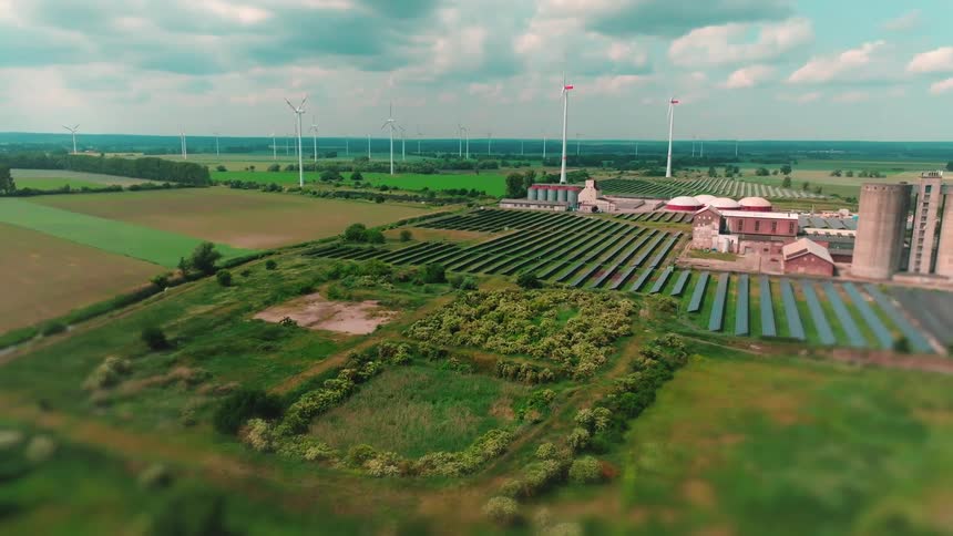The video is an aerial view of a renewable energy facility situated in a rural landscapeit shows large fields of solar panels and a number of industrial buildings all surrounded by green fields and multiple tall wind turbines in the distance