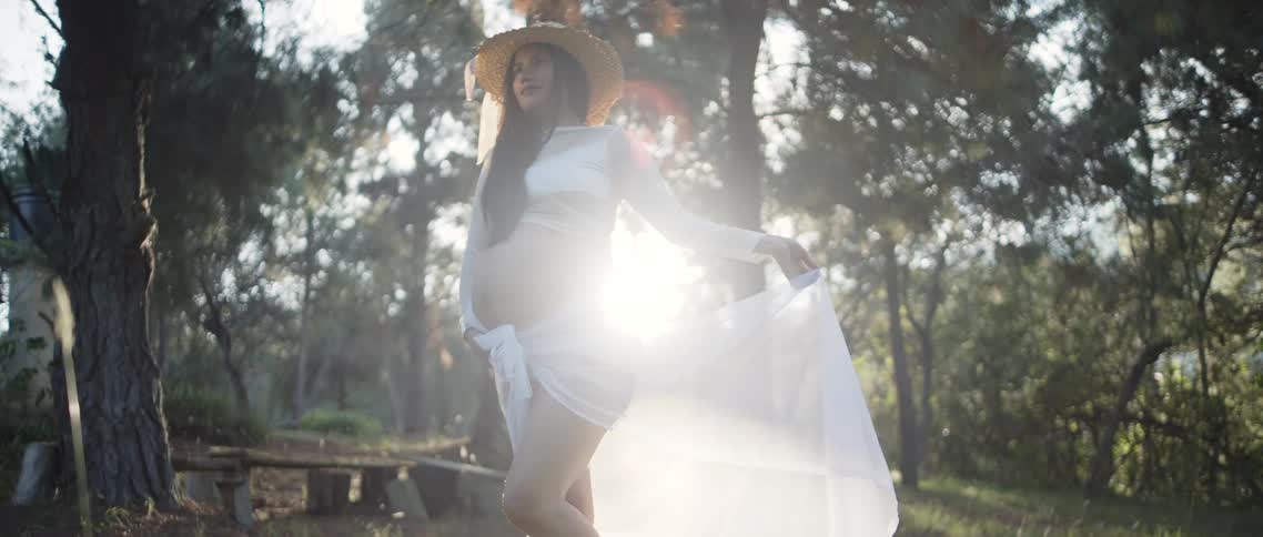 Pregnant woman wearing a hat poses outdoors holding a light fabric among sunlit trees