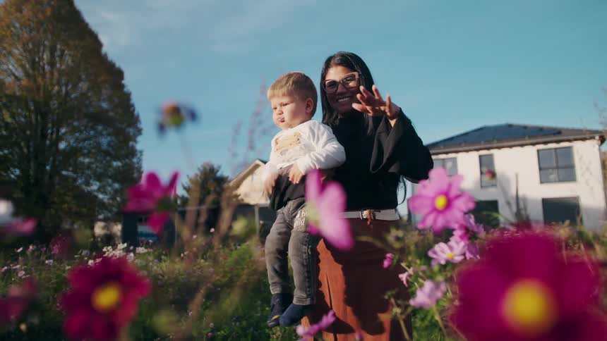 A smiling woman holding a young child waves from behind a patch of pink flowers