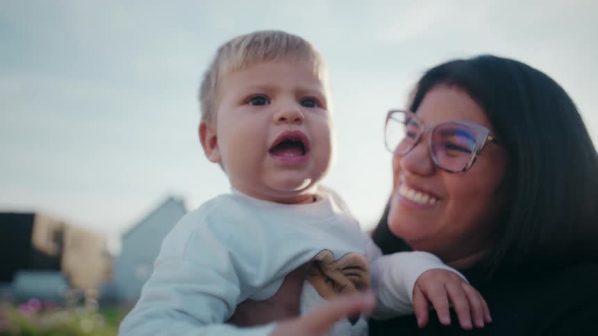 A woman wearing glasses and holding a young blondehaired child takes a closeup video or photo outdoors against a bright sky smiling warmly at the camera the child reaches out momentarily covering the lens before turning to look at and touch the woma