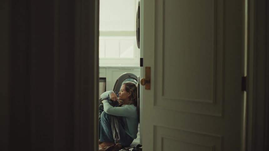 A young woman in the laundry room sorts through clothing then suddenly sits on the floor overcome with sadness
