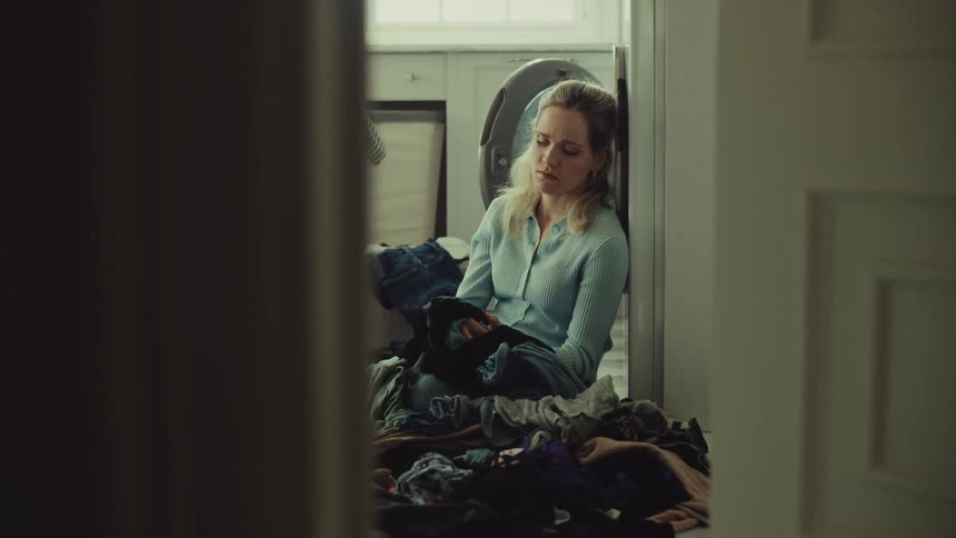 A woman is sitting on the floor of a brightly lit laundry room surrounded by a large pile of clothes she is leaning against a washing machine and holding a piece of dark clothing looking down with a somber and overwhelmed expression