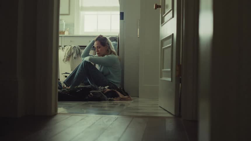 Woman sitting on the laundry room floor leans against the wall amid scattered clothes with a discouraged expression