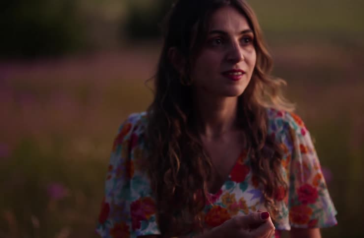 A young woman in a summer dress stands on a grassland at sunset smiling as she gazes forward