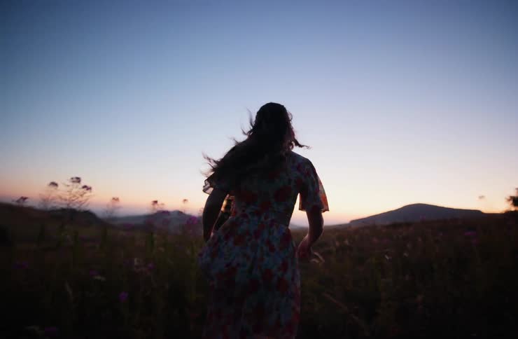 Woman walking through a flowered field at dusk toward the horizon