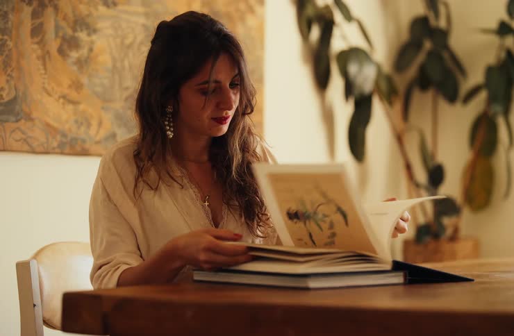 A young woman brings a stack of two books to a wooden table sits down and carefully opens the top book to examine the illustrations inside in a bright warm indoor space