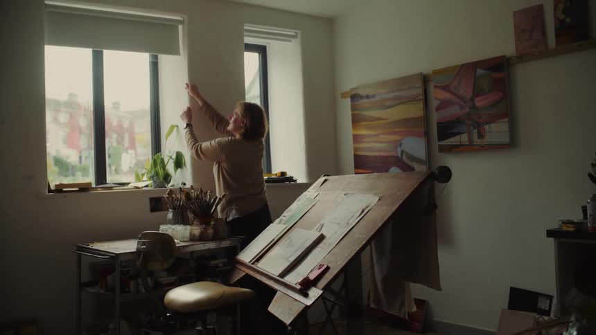 A medium shot of a woman in an artist studio raising the white window shade illuminating an easel with an art desk full of supplies nearby and paintings hanging on the wall