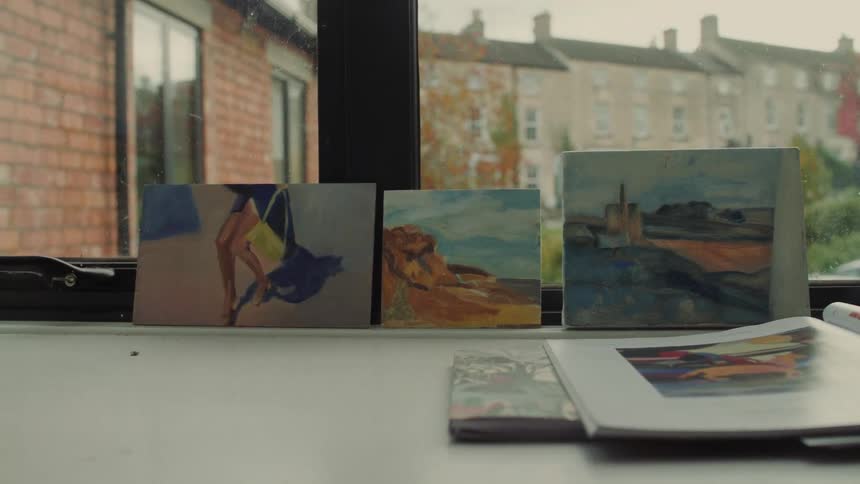 Three small colorful paintings depicting figures and landscapes are displayed upright on a window sill next to an open book with a row of brick residential buildings visible through the window pane in the background