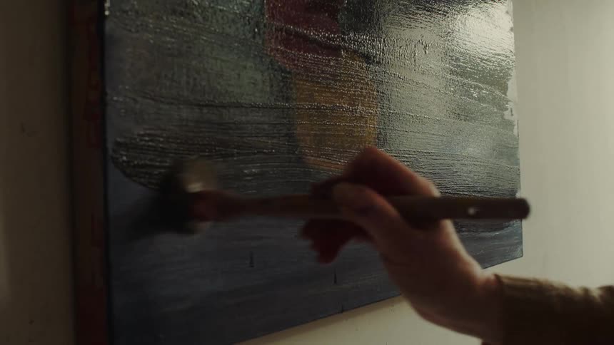 A painters hand using a wide brush to apply thick dark blue ppaint across a canvas creating horizontal brushstrokes on the surface of the artwork