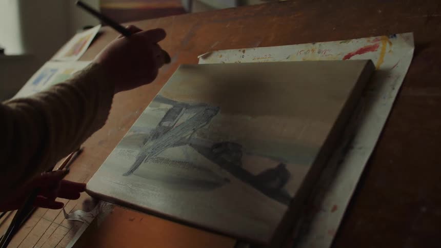 Person paints coastal scene on canvas placed on wooden surface with illustrated papers around