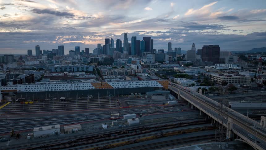 An aerial view shows a sprawling cityscape of Los Angeles dominated by numerous skyscrapers under a dramatic cloudy sky the foreground is filled with extensive railway yards and an elevated highway bridge crossing over the industrial area