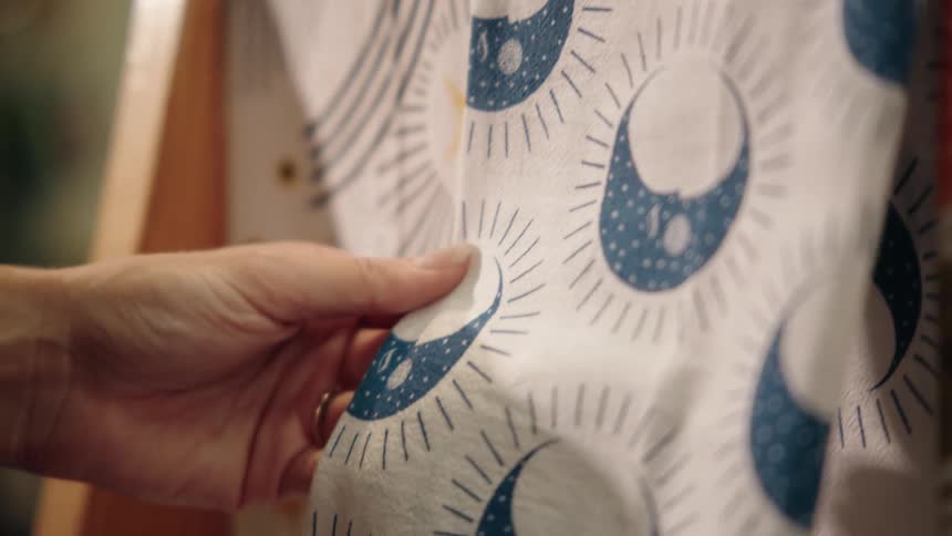 A persons hand reaches into the frame and briefly touches the white fabric of a closeup garment patterned with repeating blue crescent moon shapes and dotted rays