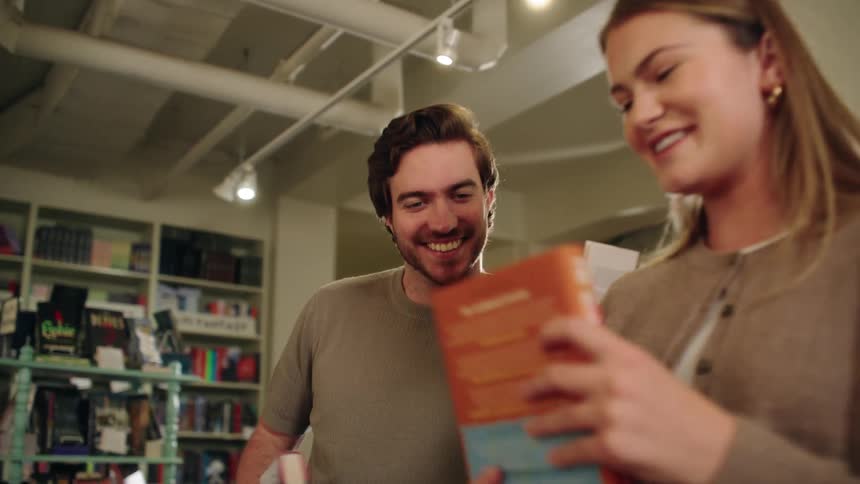 A smiling couple stands close together in a bookstore looking down at a book held by the woman while surrounding shelves full of books fill the background
