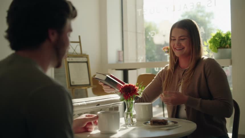 A woman with long hair wearing a cardigan smiles and gestures while talking to a man seated across from her at a small cafe table that holds two white mugs a dessert and a red flower with bright natural light coming from the window behind her