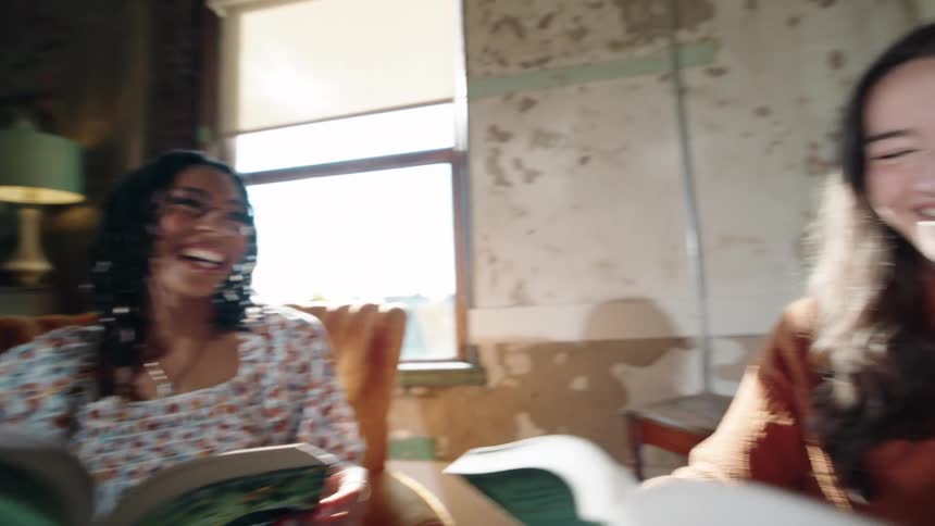Three young women are seated together each holding a book and smiling widely in a brightly lit rustic indoor space