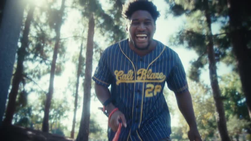 A young man wearing a blue and yellow pinstriped baseball jersey runs excitedly toward the camera in a park setting laughing while raising one hand in a gesture of celebration all brightly backlit by the sun filtering through tall pine trees