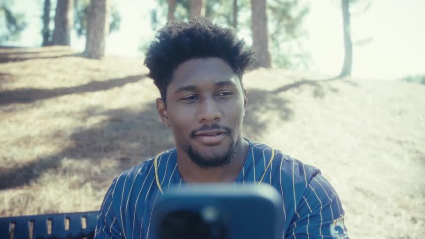 A young man sits on a bench in a public park at midday recording a selfie video