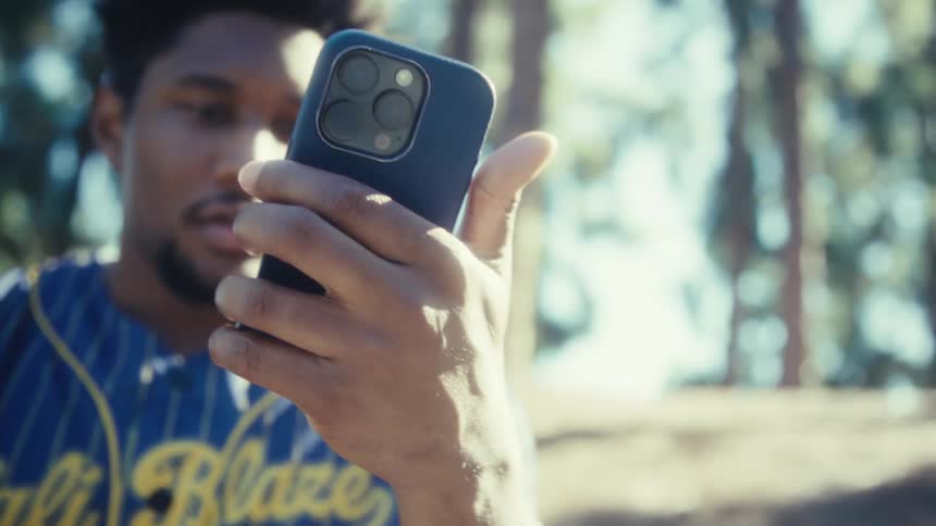 Man in sports shirt holds blue phone in front of sunlit trees