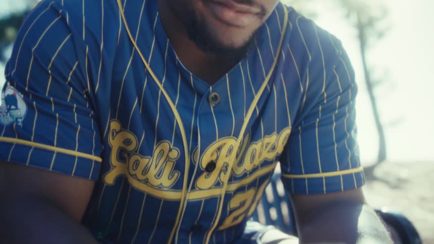 The video features a closeup shot of a young man wearing a navy blue baseball jersey with yellow pinstripes and the name cali blaze stitched across the front he is seated outdoors in a sunny slightly bright setting looking down at the smartphone he