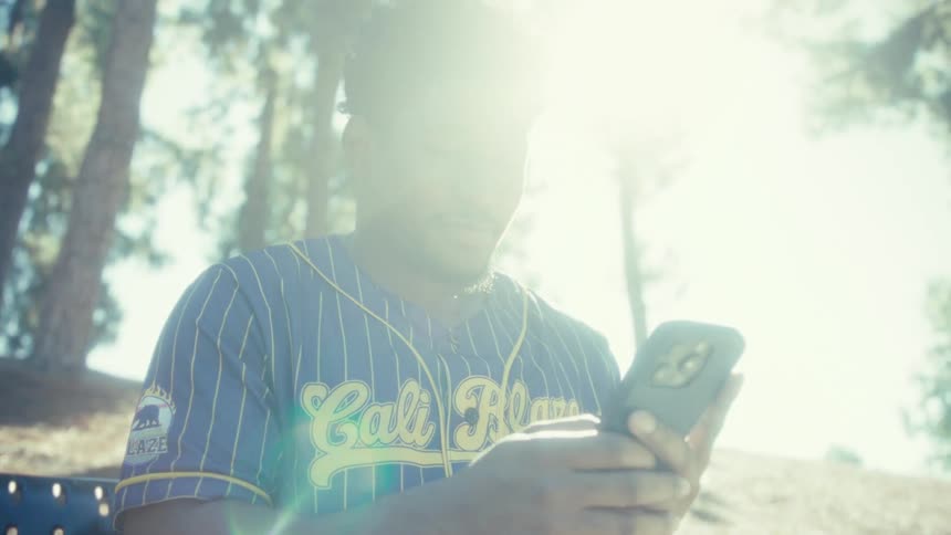 A man wearing a blue and yellow pinstripe baseball jersey is seated outdoors looking down at the smartphone in his hands with a warm smile as the bright sunlight filters through the trees behind him